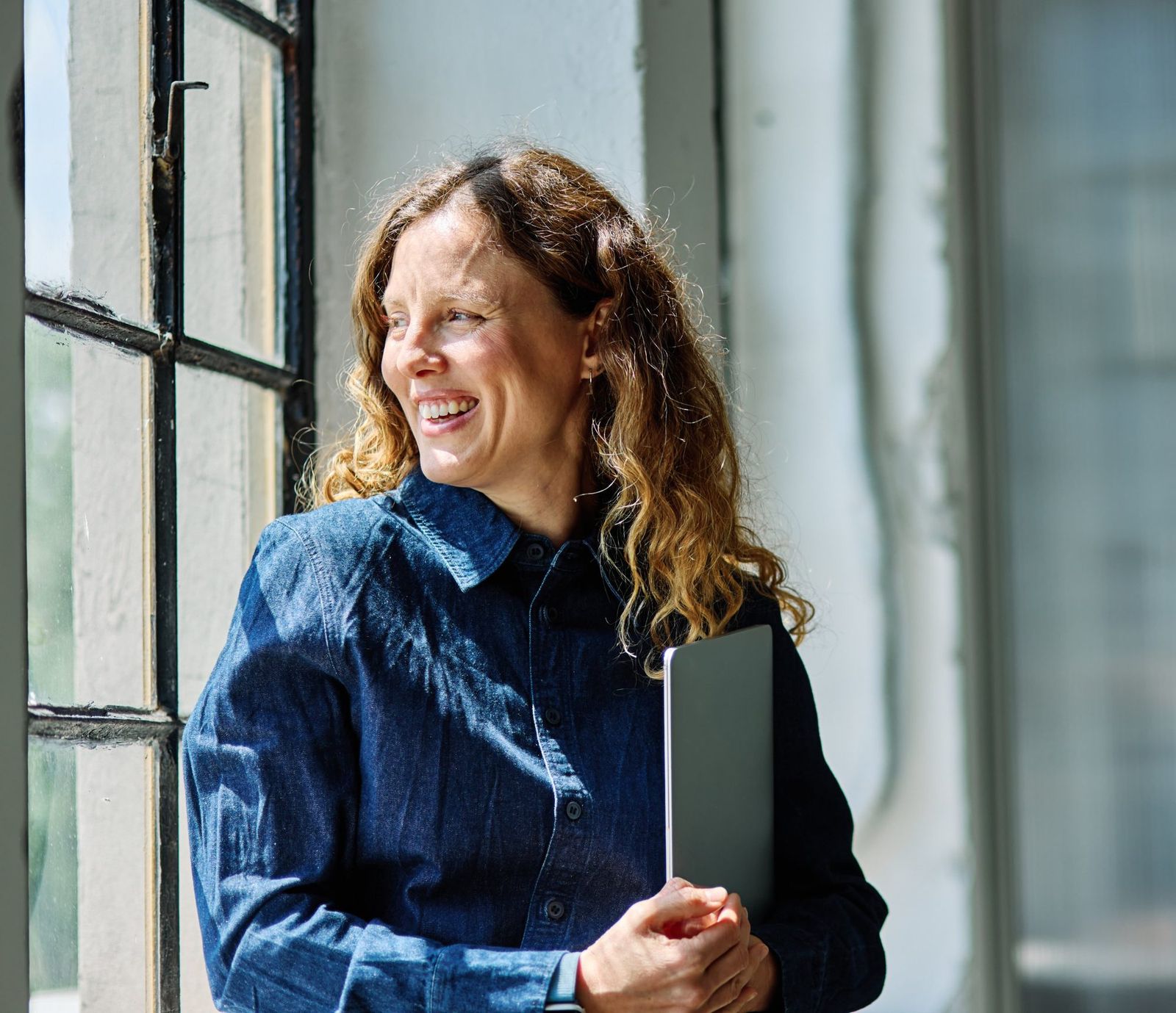 Smiling woman holding a laptop, looking out a sunlit window